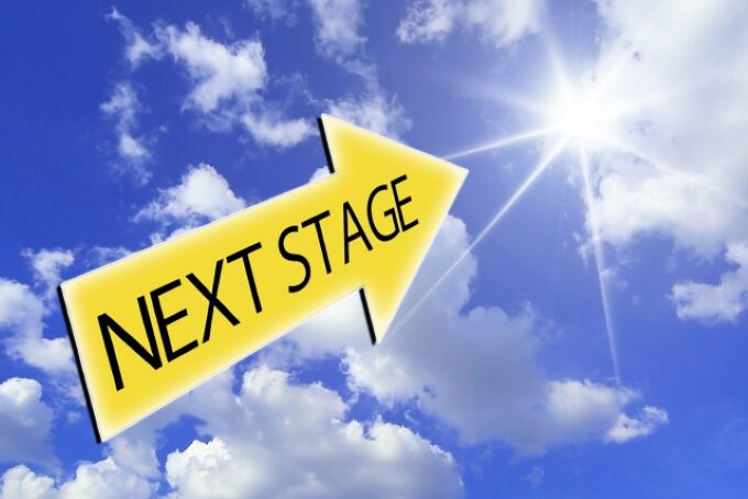 青空に向かって進む「NEXT STAGE」と書かれた黄色い矢印。次のステージや前進を象徴するイメージ。