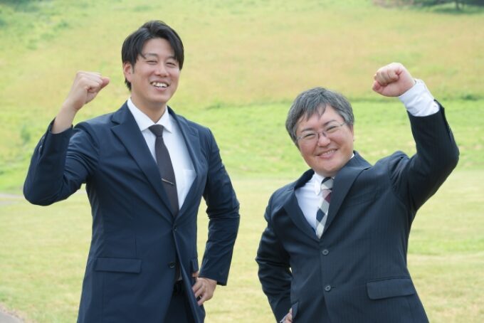 スーツ姿の男性二人が屋外でガッツポーズをしながら笑顔で並んでいる