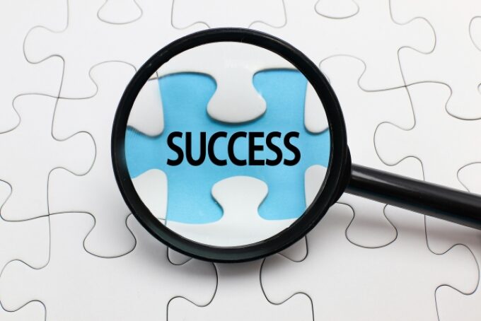 ジグソーパズルのピースに書かれた「SUCCESS」の文字を拡大鏡で強調している画像