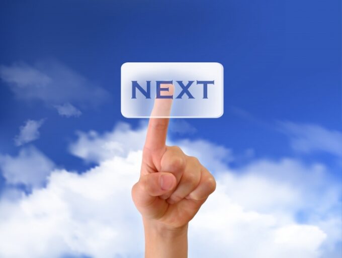 青空の下、「NEXT」ボタンを指でタッチする手の写真。次のステップへ進むイメージ