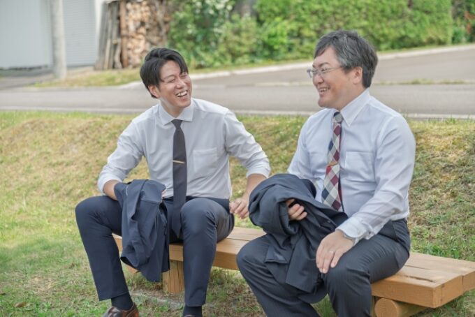 屋外のベンチでスーツ姿の男性二人が笑顔で談笑している様子