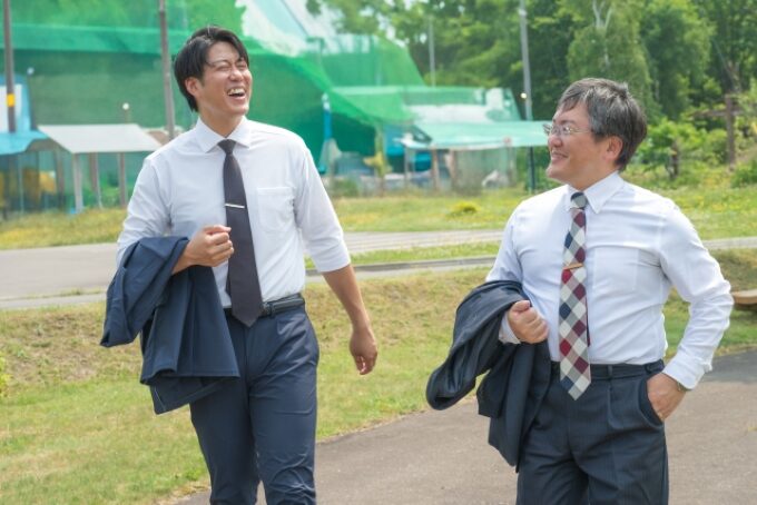 世代の異なる2人の営業マンが笑顔で歩く様子。50代と若手がともに現場で活躍するイメージを表現。